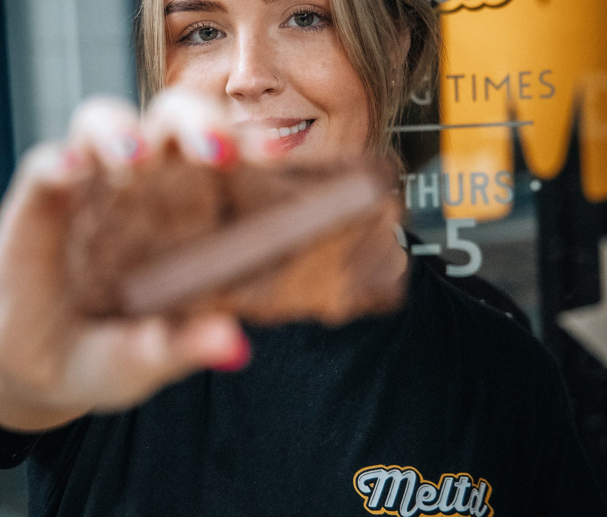 Woman wearing a black t-shirt with 'MELTD' logo in front of a store window.